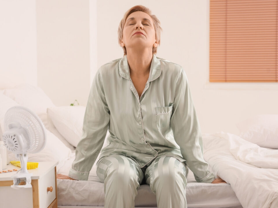 menopauza-munkahelyi-tunetek-tamogatas-x - Nők Lapja Rövid hajú nő csíkos, halványzöld pizsamában ül az ágy szélén, csukott szemmel, kissé hátrahajtott fejjel. Mellette kis ventilátor áll az éjjeliszekrényen.