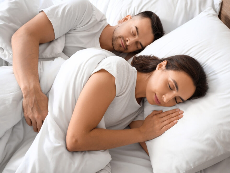 szocialis-jetlag-korabbi-lefekves - Nők Lapja Fehér pólós nő és férfi alszik egymás mellett, oldalt fekve, fehér ágyneműben; a nő kezeit összetéve a párnáján pihenteti.