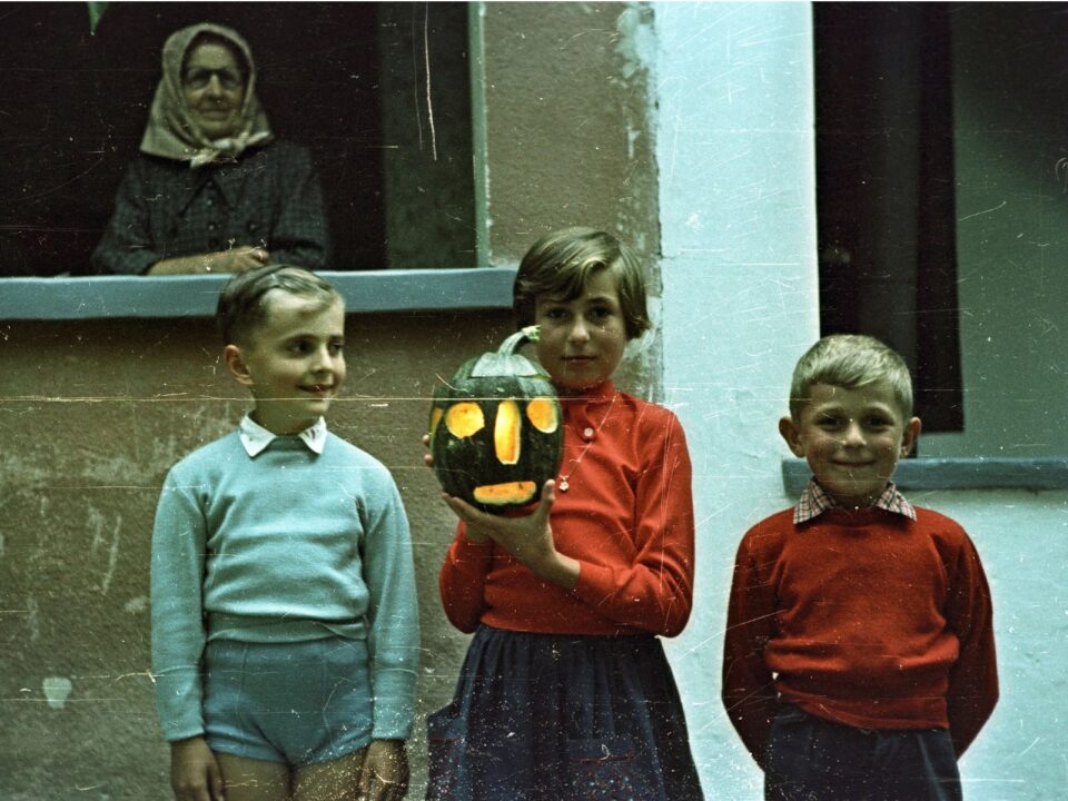 Három gyerek áll egy ház előtt Győrladaméren 1955-ben, a középen álló kislány világító, kifaragott töklámpást tart a kezében.